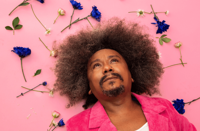 Imagem mostra homem de roupa rosa deitado em chão também rosa, com flores ao redor da sua cabeça