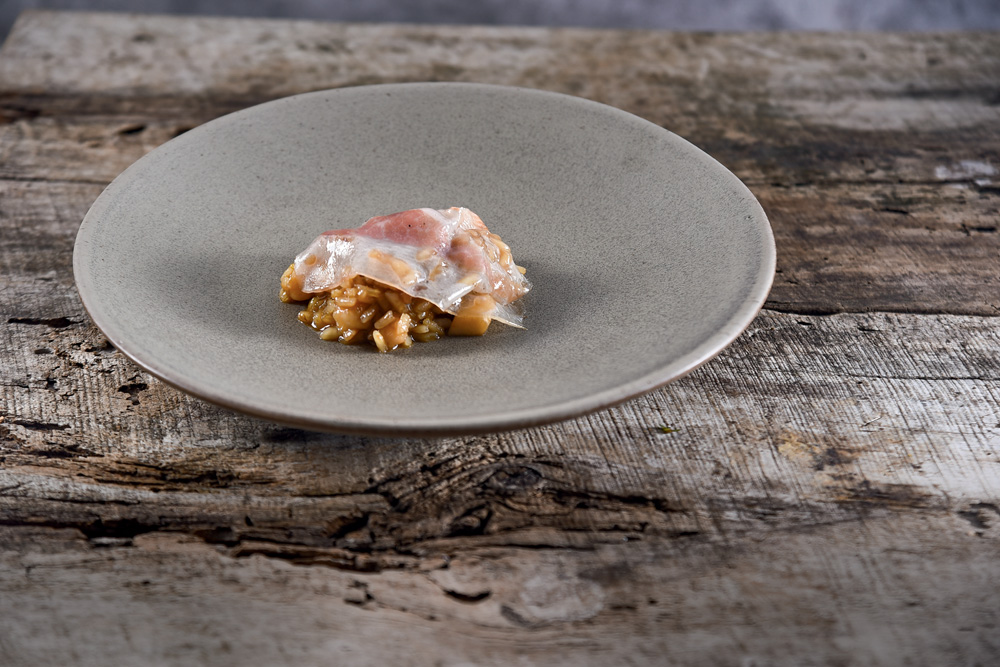 Prato de cerâmica cinza sobre mesa de madeira contendo arroz de garoupa com uma lâmina de guanciale