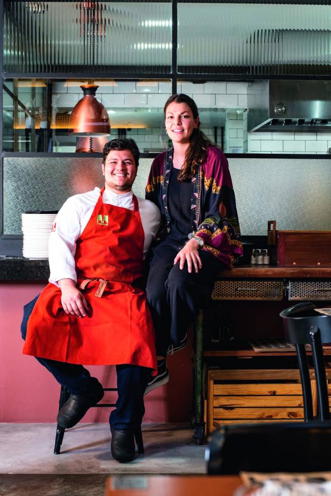 Casal com homem vestindo avental vermelho e sentado sobre balcão em ambiente do restaurante