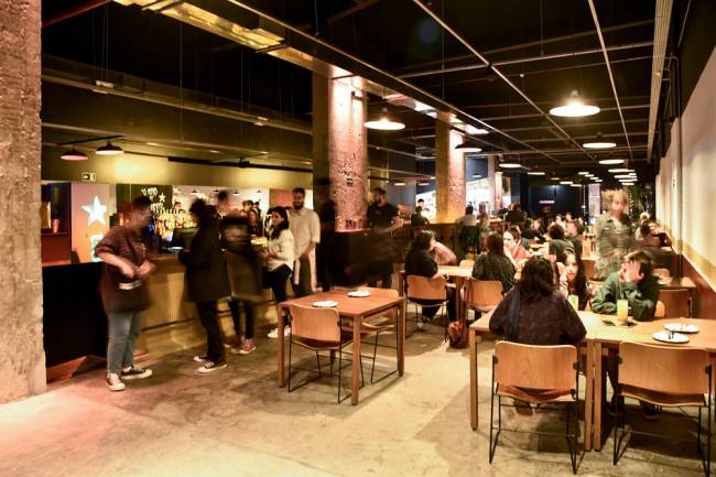 Ambiente interno de bar, com pessoas em pé e sentadas à mesa