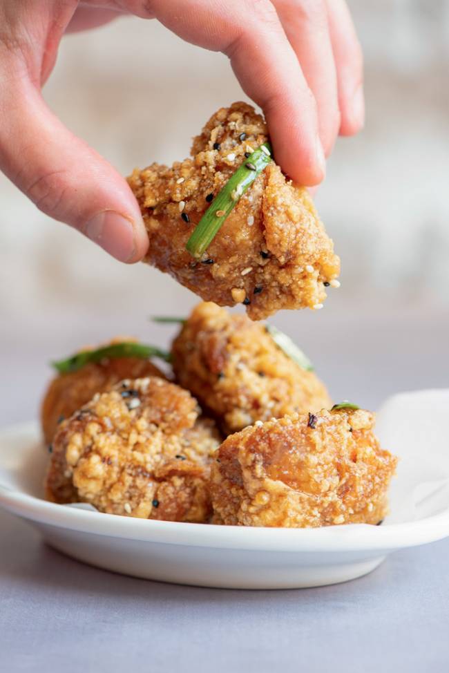 Frango frito, chinkiang vinagrete gergelim nirá – Mila Prato de porcelana com pedaços de frango frito, um dos pedaços sendo segurado por uma mão de pele branca
