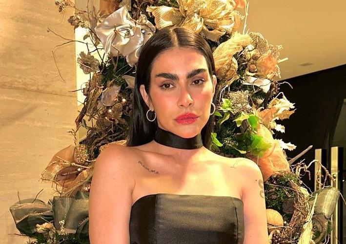 Foto mostra Cleo com sobrancelhas laminadas, semblante neutro, vestido preto, ao fundo árvore de natal