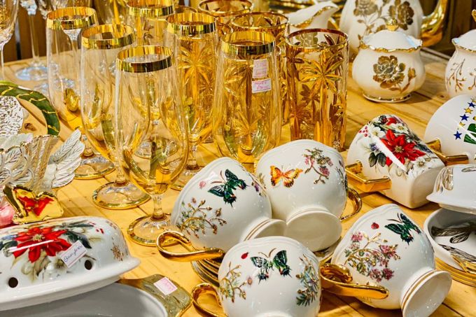 Xícaras de porcelana com desenhos de borboletas e flores. Ao fundo, há copos de vidro com detalhes em ouro. Todos os objetos estão dispostos em uma mesa