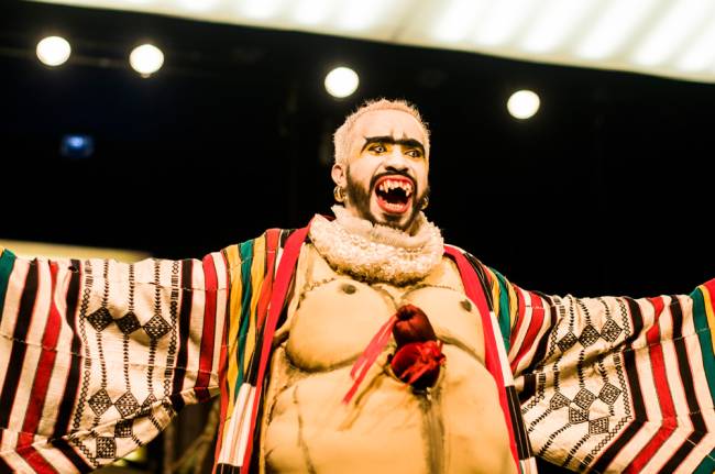 Ubu Rei O ator Douglas Novais estende os braços e sorri no palco. Ele usa lentes de contato chamativas, dentes afiados e figurino bizarro e colorido