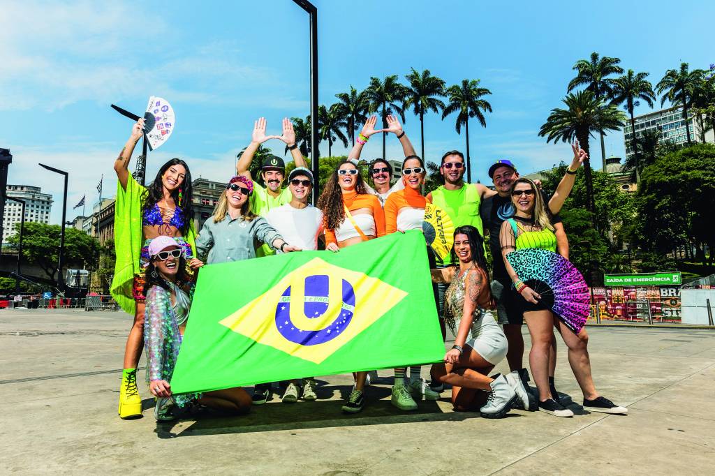 Ultra Brasil Imagem mostra grupo de pessoas vestindo roupas de carnaval, alegres, e segurando bandeiras e cartazes de festival de música. Ao fundo, um céu azul.