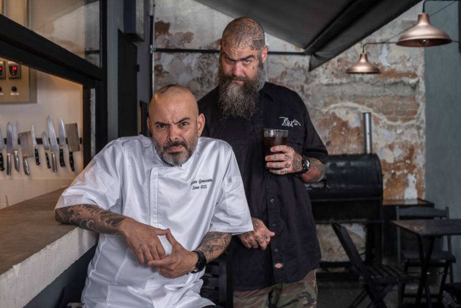 Um homem careca de camiseta branca de mangas curtas e homem também careca de barba longa com camiseta preta, ambos tatuados, olham para a câmera de forma descontraída, fazendo careta