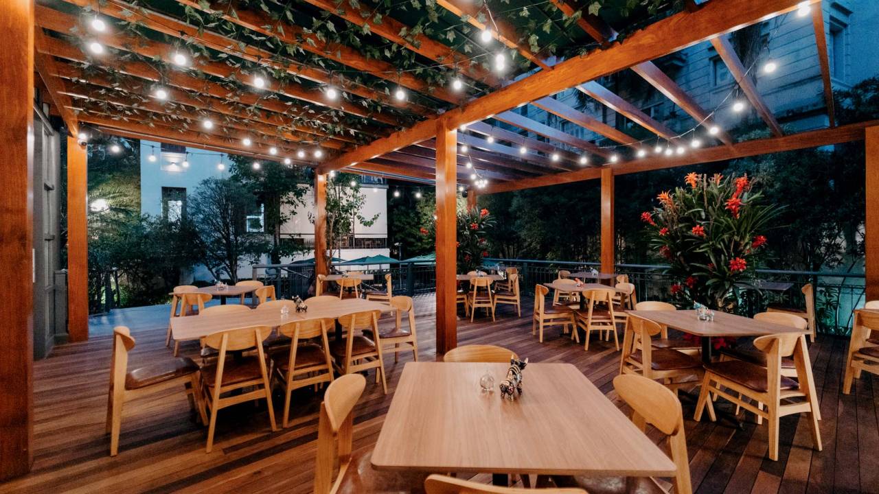 Espaço de restaurnate sob marquise com plantas, em chão elevado de madeira, com mesas de madeira clara e cadeiras amarelas