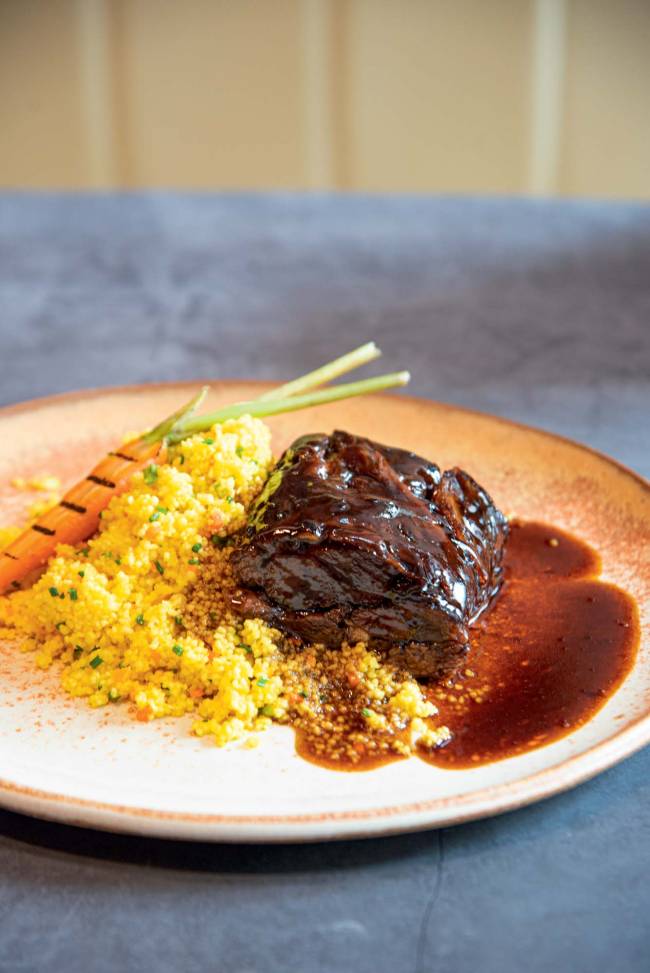 Paleta de cordeiro ao molho do assado e cuscuz marroquino – Casa La Pastina Prato de cerâmica sobre mesa azul, com paleta de cordeiro, cuscuz marroquino temperado e uma minicenoura com marcas de grelha