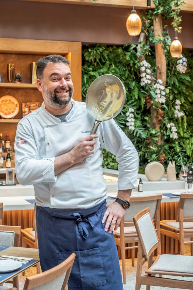 Douglas Benatti – Casa La Pastina Douglas Benatti, homem de cerca de 50 anos, com dolma e avental azul, segura frigideira em frente a parede com flores e estante, além de haver mesas e cadeiras de madeira clara ao fundo