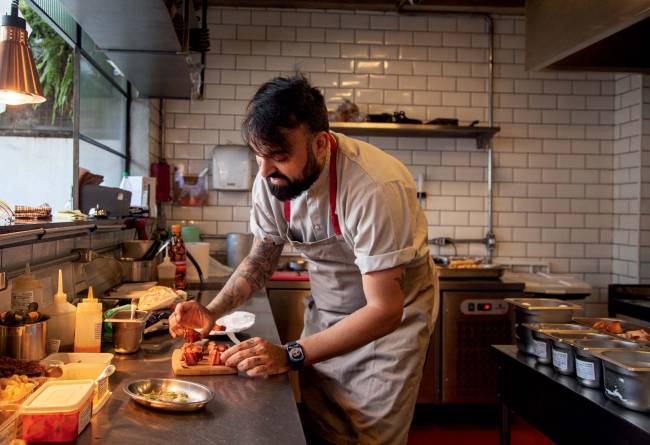 Chef diego gimenez se debruça sobre bancada com camisa e avental beges e finaliza prato