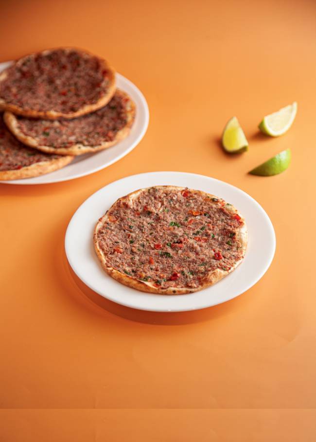 pilha de esfirras abertas de carne sobre pratos em cima de mesa laranja, com limão cortado em cunhas ao lado, sobre a mesa