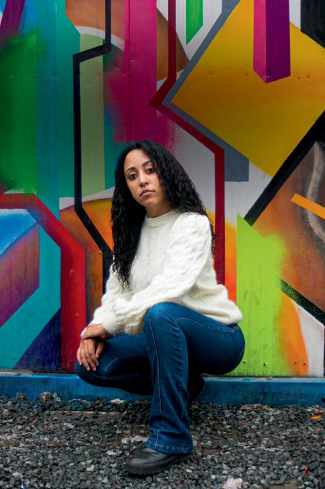 Ester Carro Mulher abaixada em frente a muro grafitado
