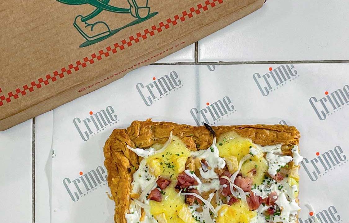 Pedaço quadrado de pizza sobre papel manteiga onde se vê o logo da Crime Pastry Shop
