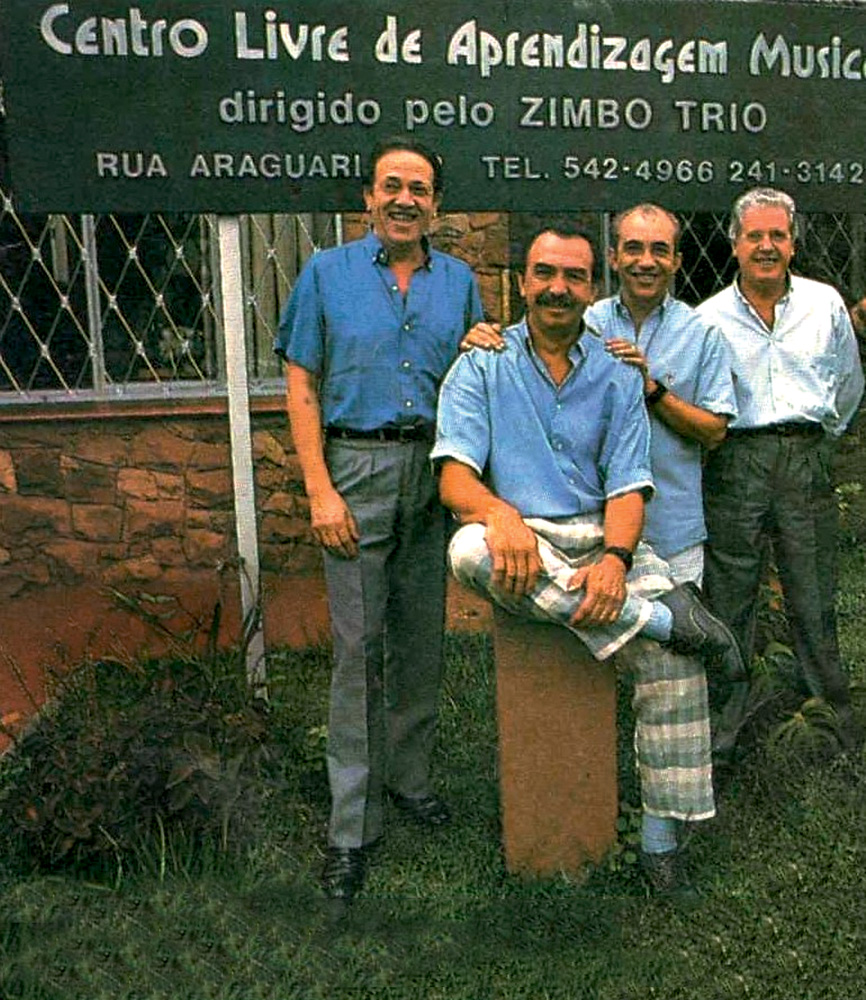 Clam 50 anos Imagem mostra quatro homens de camisa social sorrindo, ao lado de placa de escola