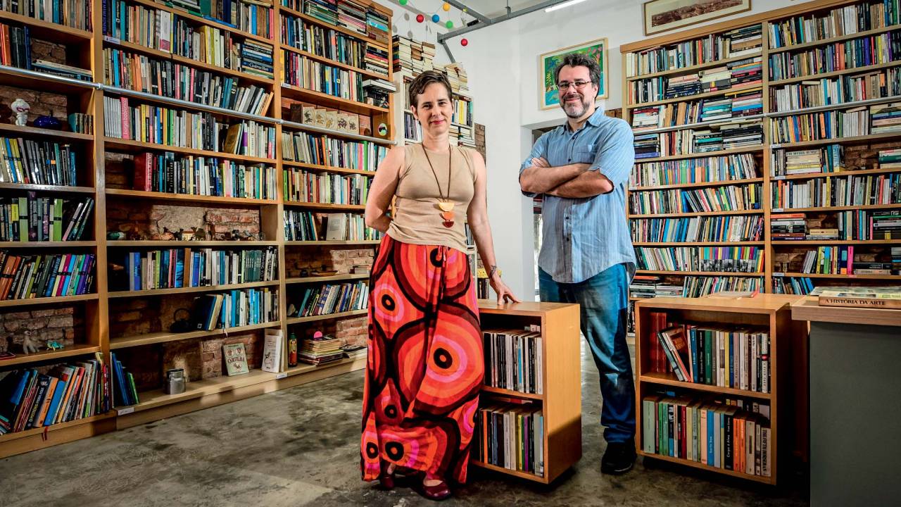 Imagem mostra sala com estantes de livros e uma mulher e um homem, ao centro, sorrindo