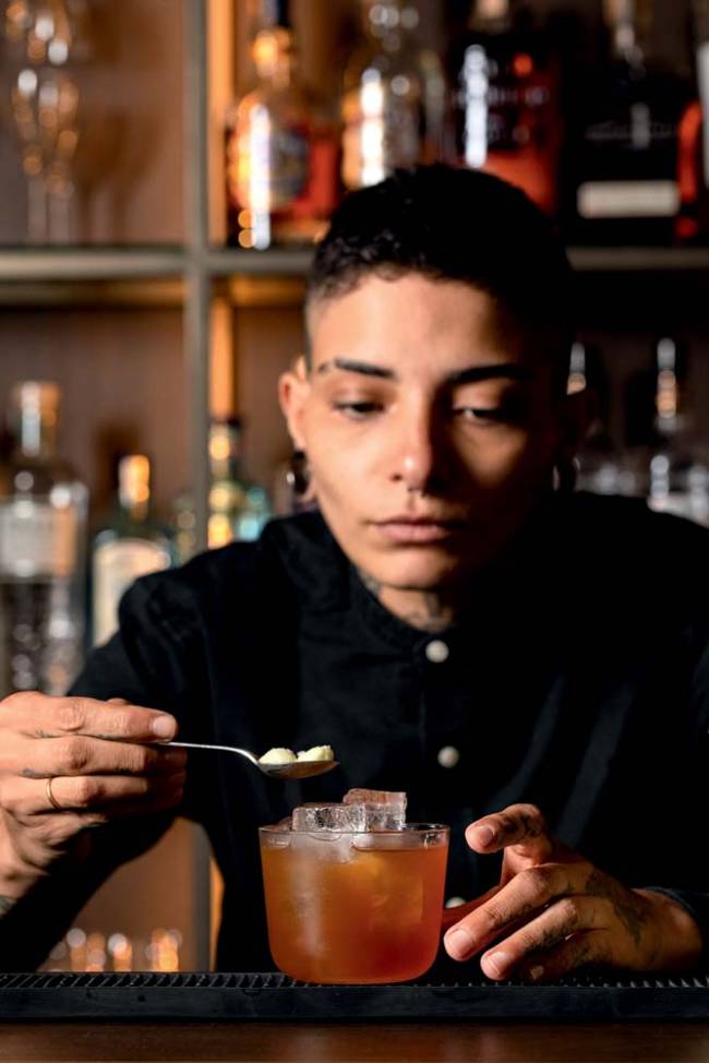 Mulher de cabelo curto, raspado à máquina, segura colher como se adicionasse algo a um drinque de copo baixo alaranjado que está à sua frente