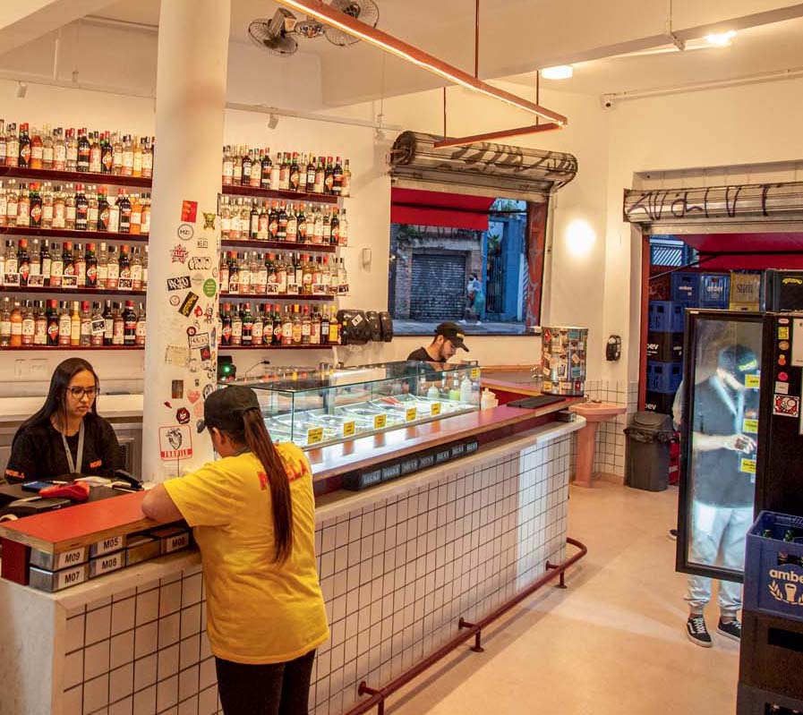 Moela Imagem mostra interior de bar, em branco e vermelho, com uma estante de bebidas na parede e alguns clientes sentados