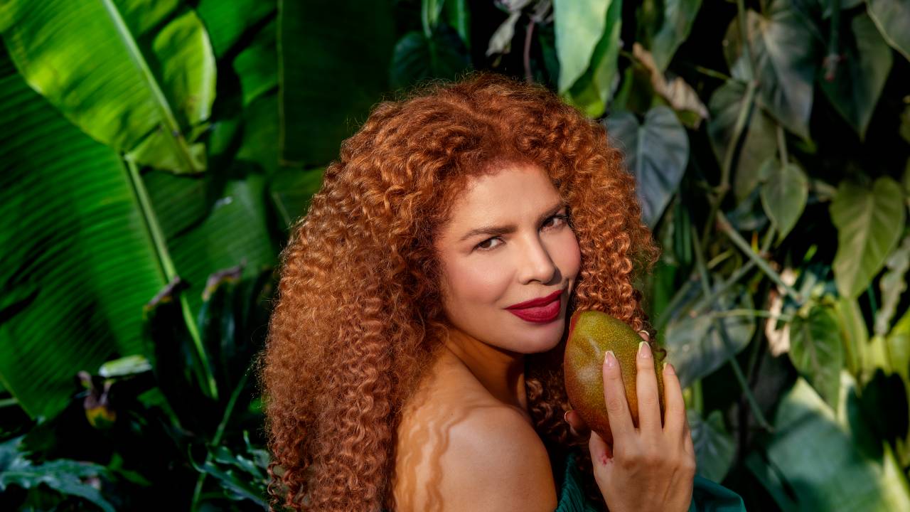 Imagem mostra mulher de cabelo ondulado segurando manga, com floresta ao fundo