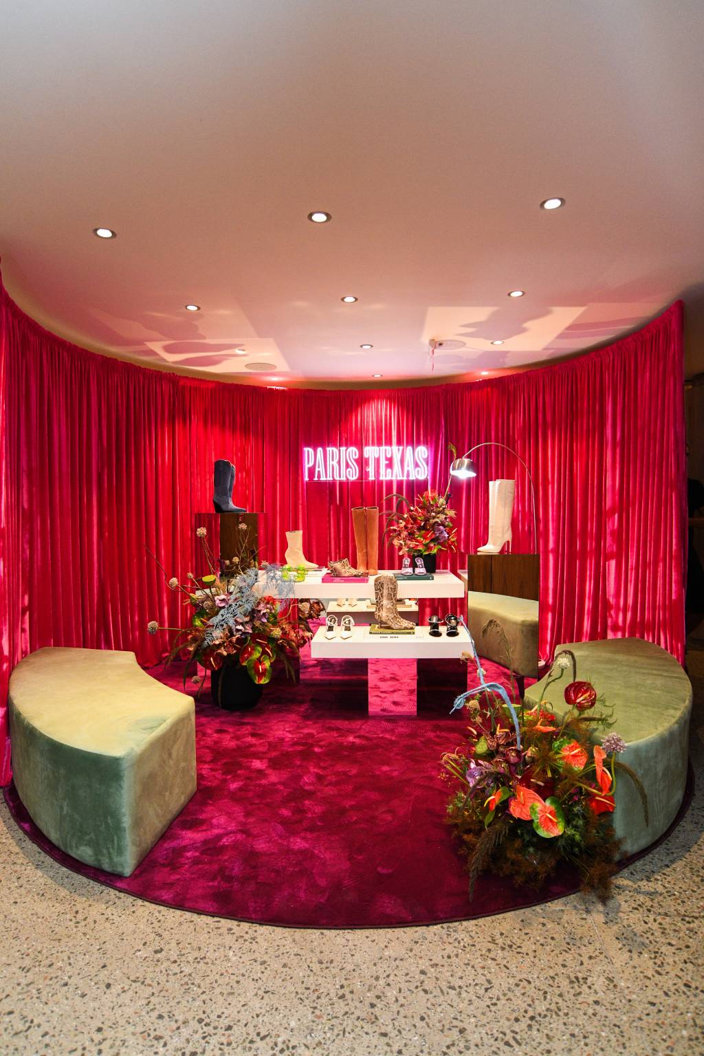 Paris Texas na NK Store. Neon rosa 'Paris Texas' aparece em destaque em frente a cortina de veludo e ilumina algumas bancadas claras e repletas de sapatos e botas.