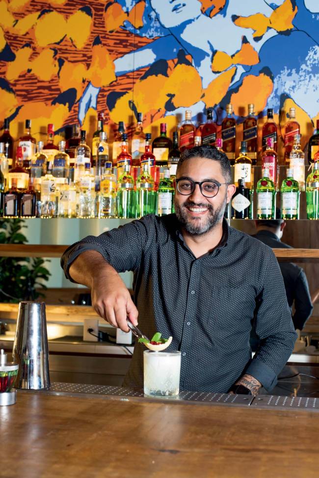 Ricardo Barrero Bartender Ella Fitz Homem de camisa preta prepara drinque sobre bancada de madeira, com garrafas no bar ao fundo e ao lado de coqueteleira prateada