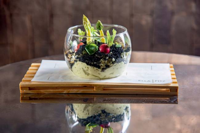Terrario Legumes Ella Fit Recipiente de vidro em formato de terrário contendo legumes e espuma de aspargos verde que lembra a areia colorida dos terrários de verdade