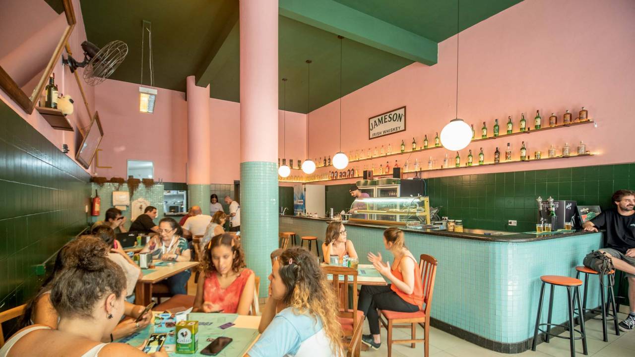 Ambiente interno do escarcéu com paredes cor-de-rosa, cobertas na metade inferior com azulejos verdes, com mesas de madeira e clientes sentados
