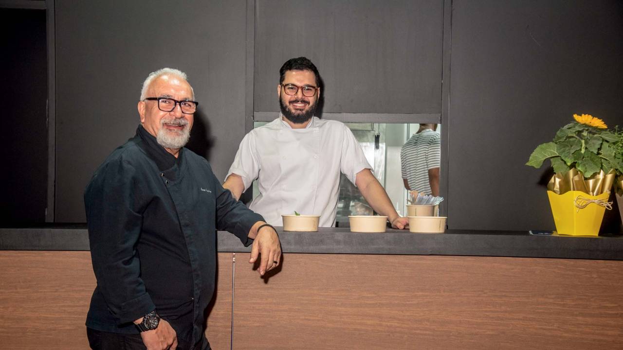 Homem de dolma atrás de balcão e outro na frente, grisalho, vestindo preto