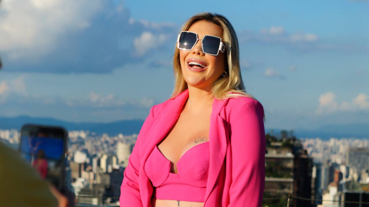 Mulher branca e loira posa no topo de prédio com roupas rosa pink e bolsa na mão direita.