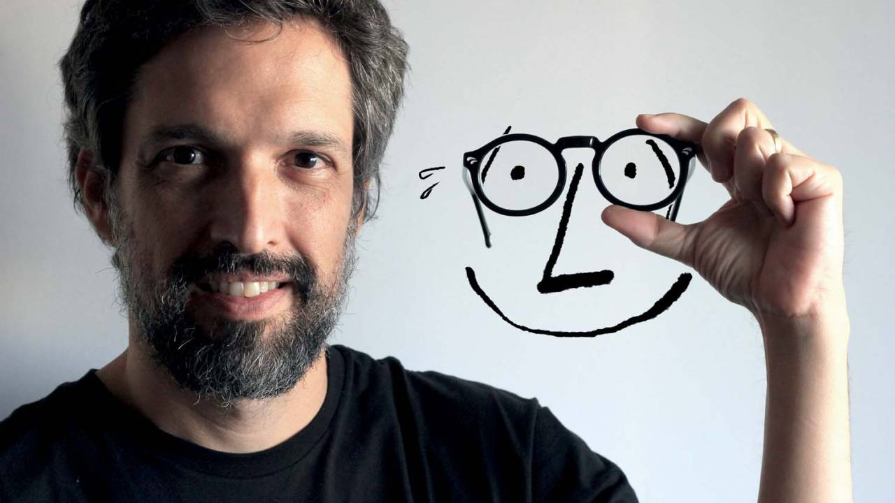 Homem branco de barba grisalha sorri segurando traços de um personagem com óculos e cara de preocupação.