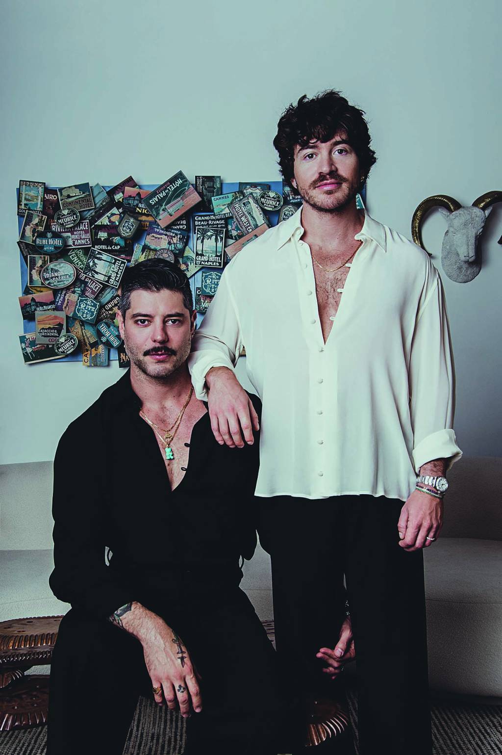 Kadu Dantas e Peter Zawel (Carlos Neto-Divulgação).tiff (1) Dois homens brancos encaram a câmera, o da esquerda sentado com topete, bigode e camisa preta. O da direita de pé, apoiando o braço no ombro do outr, com cabelos escuros, barba e camisa branca com botões abertos no peito.