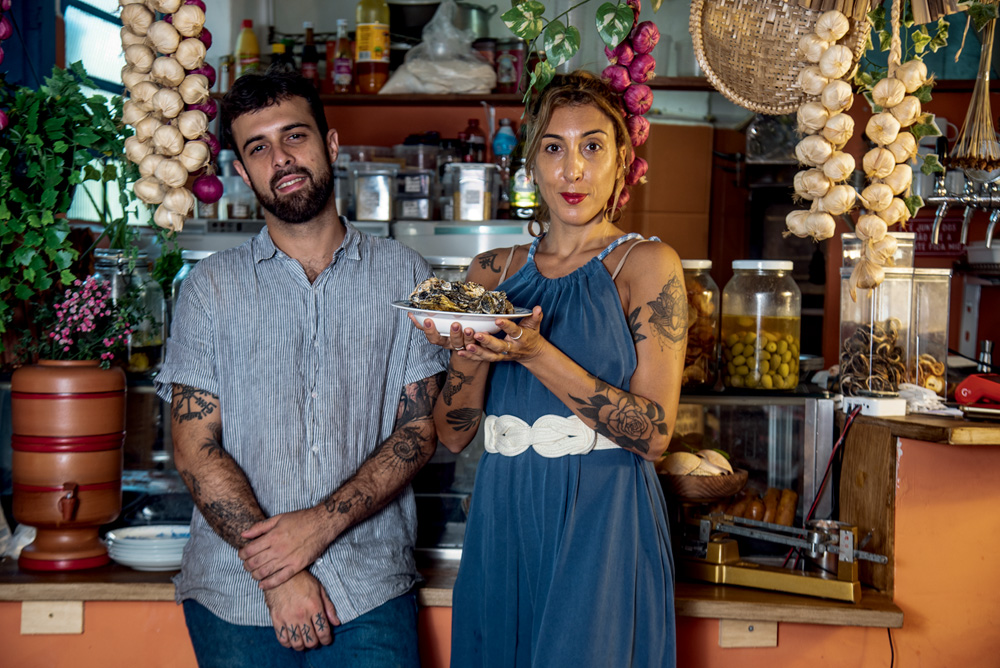 Andrea Verissimo e Bruno Mendonça – Manu. el a Tasca Homem e mulher brancos de braços tatuados a frente do balcão alaranjado do bar Manu.el.