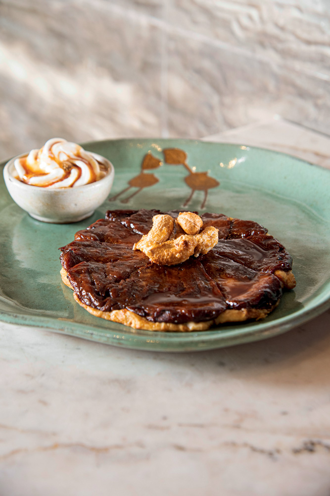 Prato de cer&acirc;mica verde com torta doce achatada coberta por castanha-de-caju caramelada ao lado de potinho com sorvete.
