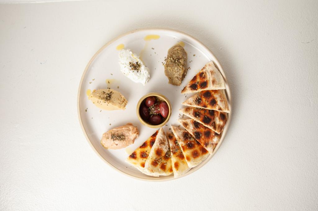 Petros Greek Taverna Foto aérea de mézzes, prato branco em cima de tampo branco com quatro pastas gregas de um lado, um potinho de azeitona ao centro e pães pita cortados em triângulo à direita.