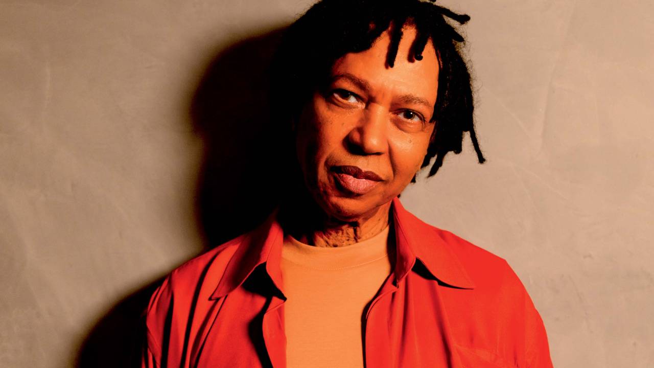 Cantor Djavan em fundo rosa, usando uma camisa laranja