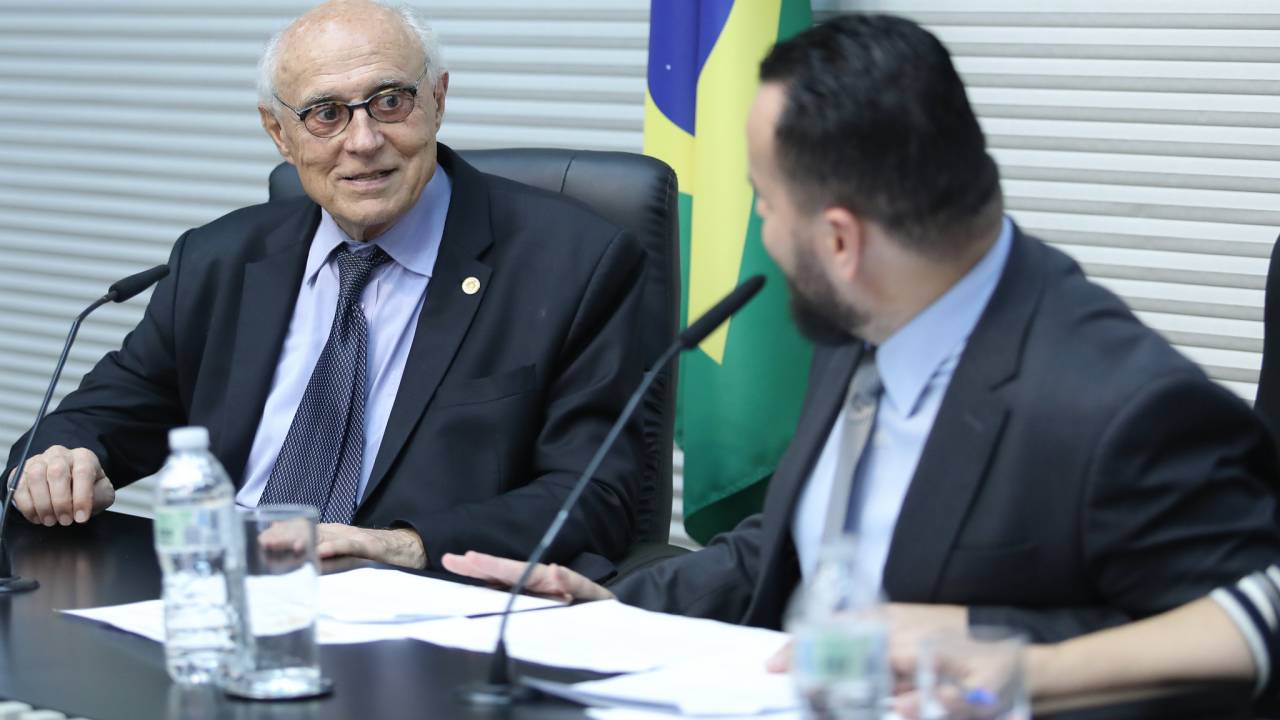Um homem grisalho conversa com um homem de cabelo castanho em uma mesa. Atrás, a bandeira do Brasil.