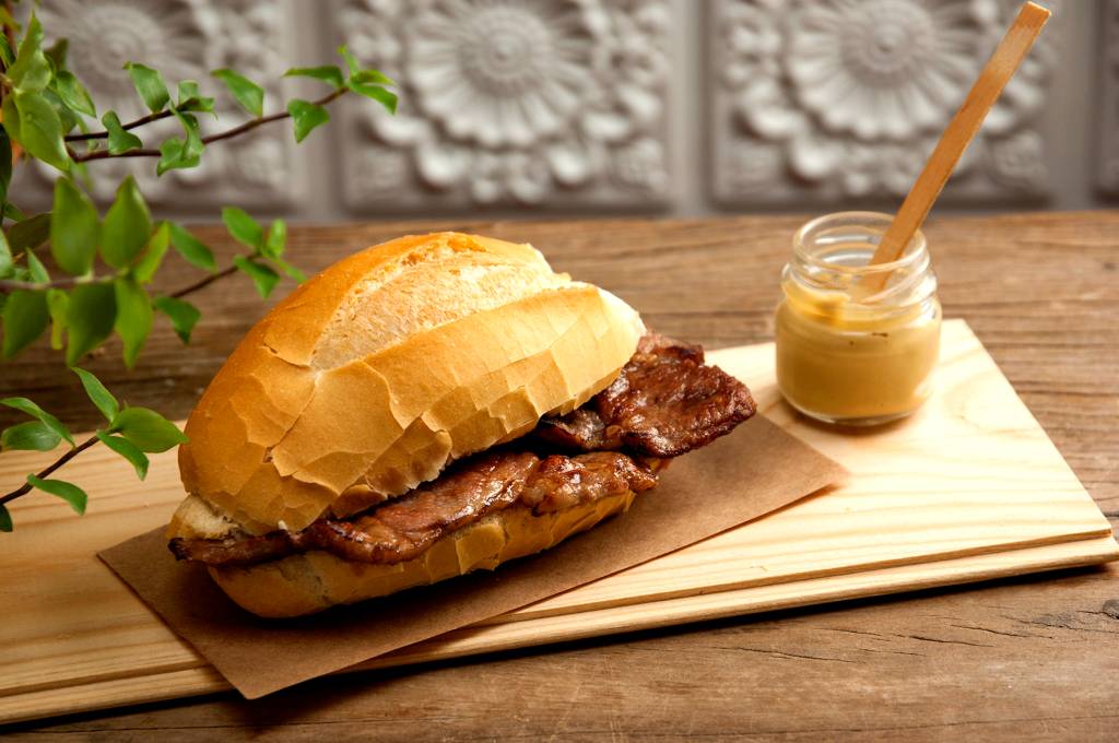 Tasca da Esquina – Bifanas Sanduíche no pão francês sobre papel krafr com pedaço de carne aparecendo ao lado de potinho de maionese.