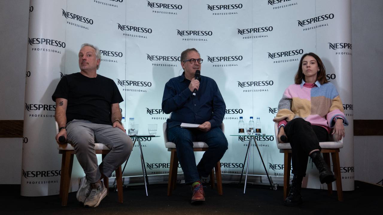 Dois homens e uma mulher brancos sentados em palco com fundo da Nespresso.