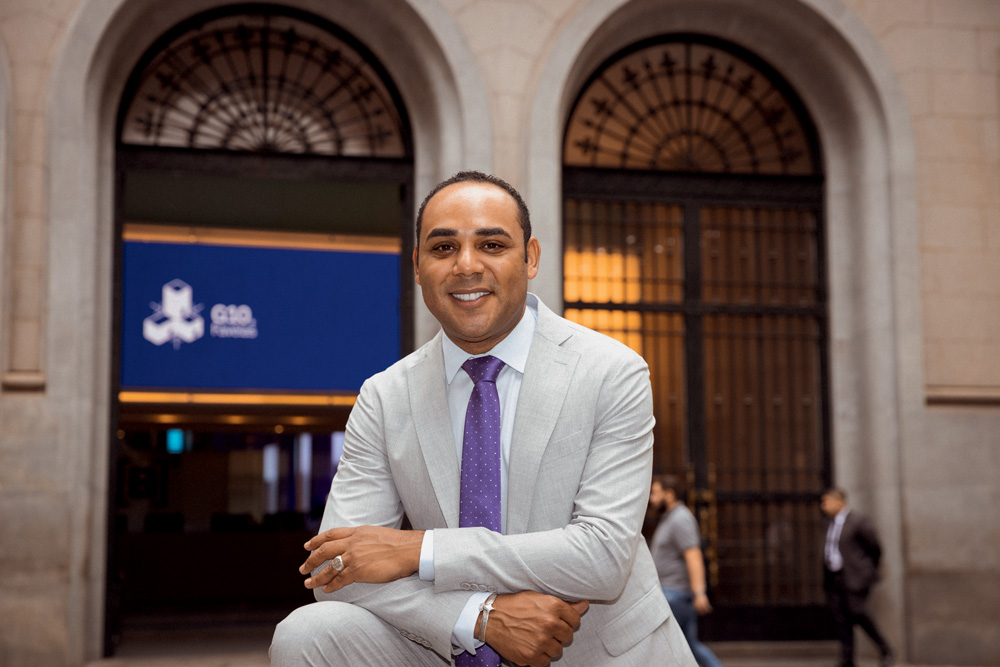 Homem negro posa de terno cinza claro e gravata roxa com os braços cruzados, sorrindo.