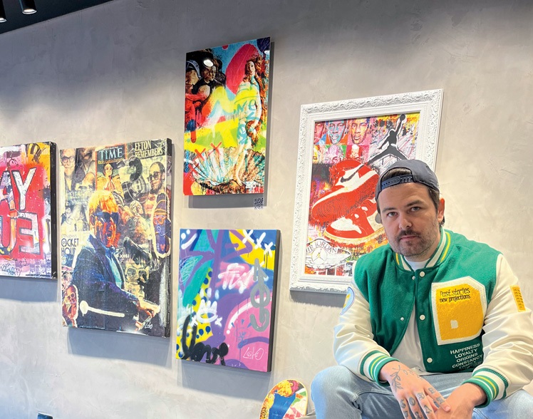 Homem branco posa em cima de poltrona de couro preto, com quadros coloridos à sua esquerda. Veste camisa verde e branca com jeans e tênis.