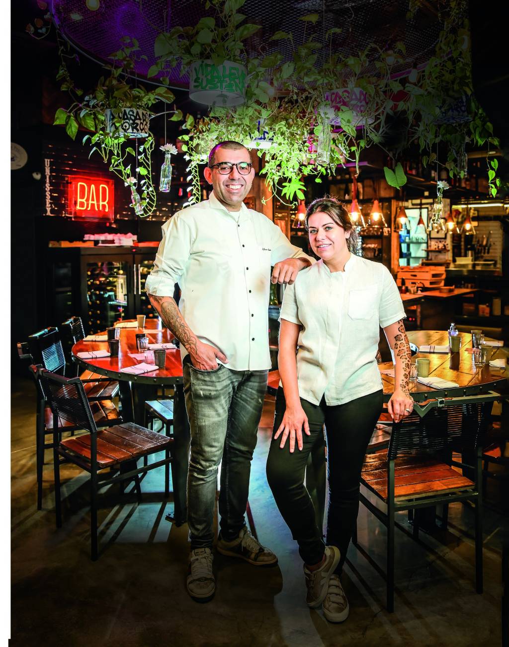 Janaina e Jefferson Rueda Casal branco de chefs trajados de dólmã no meio do salão de restaurante.