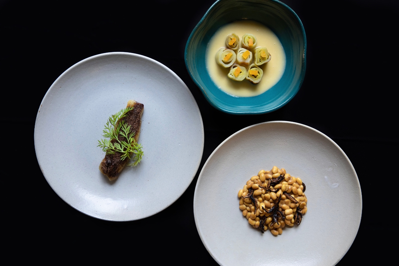 Foto a&eacute;rea de tr&ecirc;s pratos. &Agrave; esquerda, prato de lou&ccedil;a branca com fatia ao centro de berinjela assada. No canto superior direito, cuia de cer&acirc;mica azul com peda&ccedil;os de alho-por&oacute; em caldo. No canto inferior direito, prato de lou&ccedil;a branca com cozido de soja.