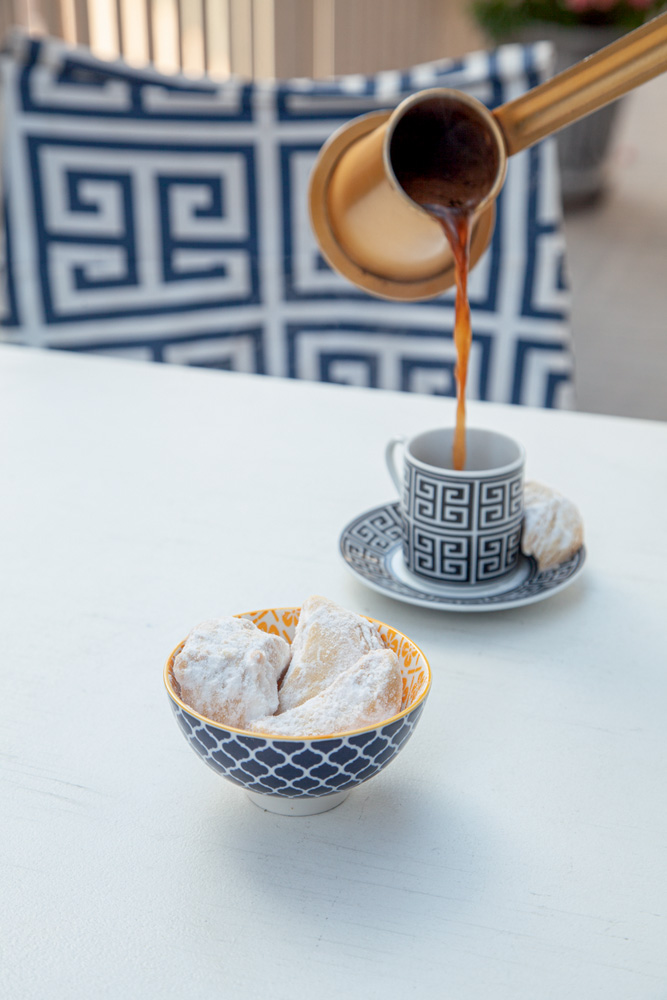 Prato Grego Café sendo servido de jarra de cobre ao fundo e cumbuca de louça azul com biscoitos envoltos em açúcar.