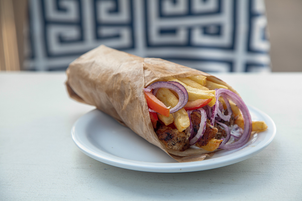 Prato Grego Espécie de kebab sobre prato de louça branca com frango, cebola-roxa e batata frita.