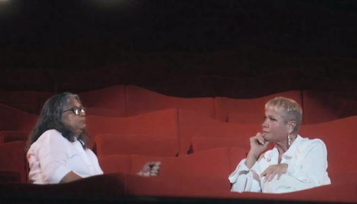 Marlene Mattos e Xuxa sentadas uma de frente pra outra em cadeiras de um teatro