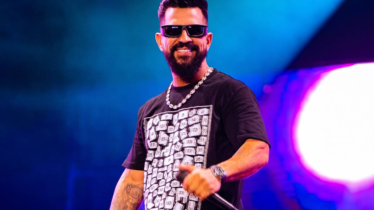 Cantor com camiseta preta e óculos escuros aparece em palco de pé com microfone na mão.