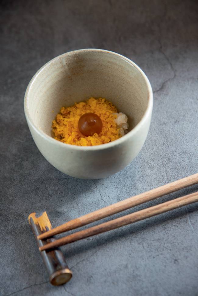 Cumbuca de cerâmica com ova de bagre ao lado de hashi apoiado em fundo cinza.