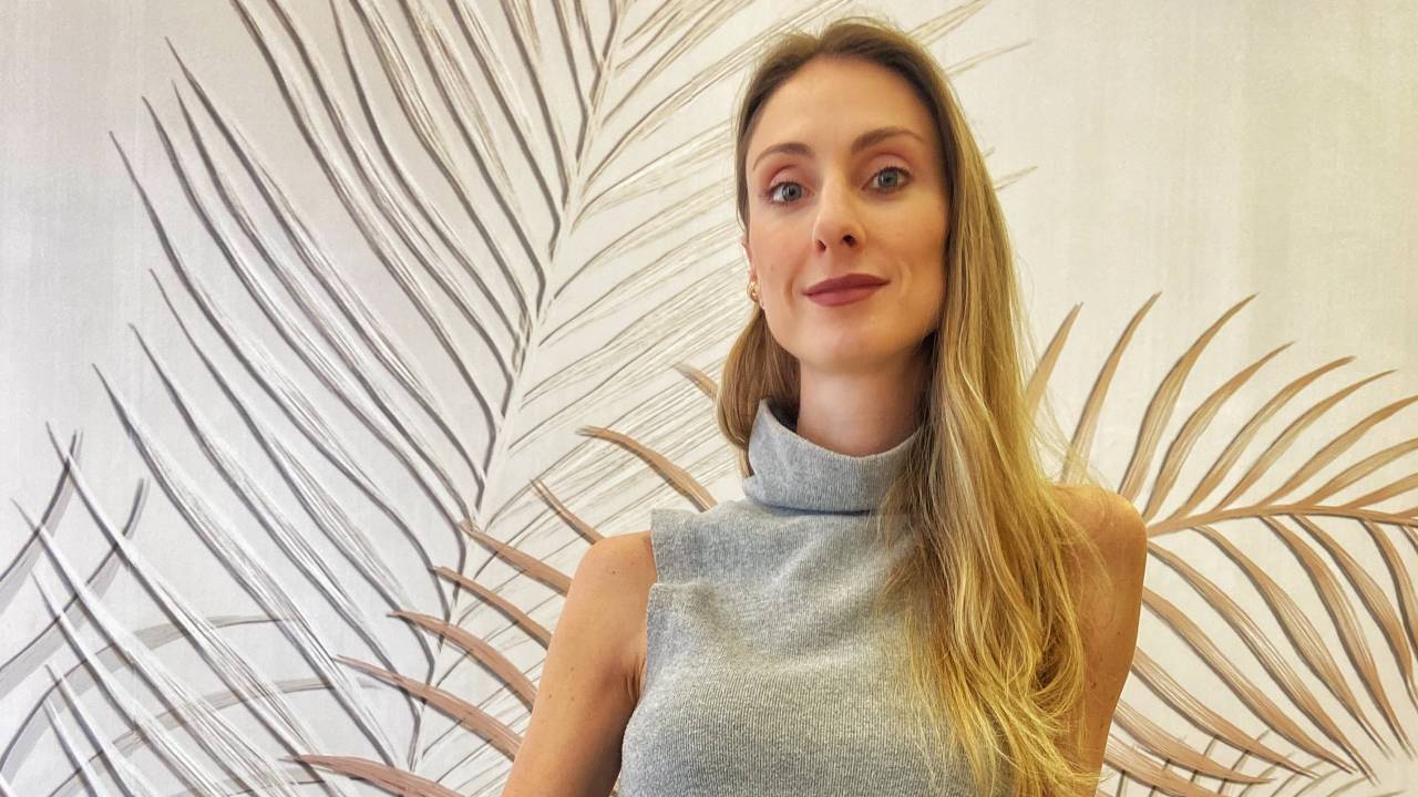 Mulher loira posa apoiada em mesa com produtos cosméticos atrás. Veste blusa de seda cinza claro e saia preta.