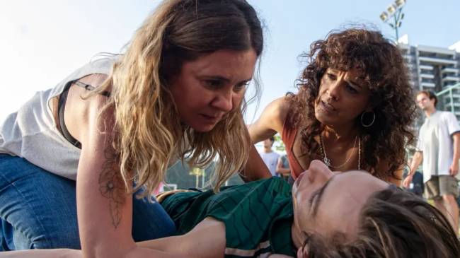 Os outros – Globoplay Menino caído no show, enquanto uma mulher branco, de braço tatuado, está agachada com as mãos no menino, enquanto uma mulher branca, de cabelos cacheados, está ajoelhada olhando em direção a outra mulher.