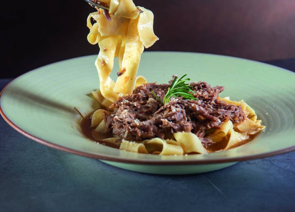 Prato fundo de cerâmica com pappardelle sob ragu de carne com garfada sendo levantada.