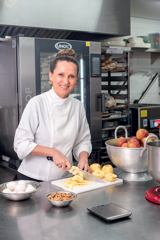 A Casa de Antonia Chef de cozinha loira com cabelo preso em coque e vestida de dólmã cortando pêssegos em cozinha industrial de restaurante ao sorrir para a foto.